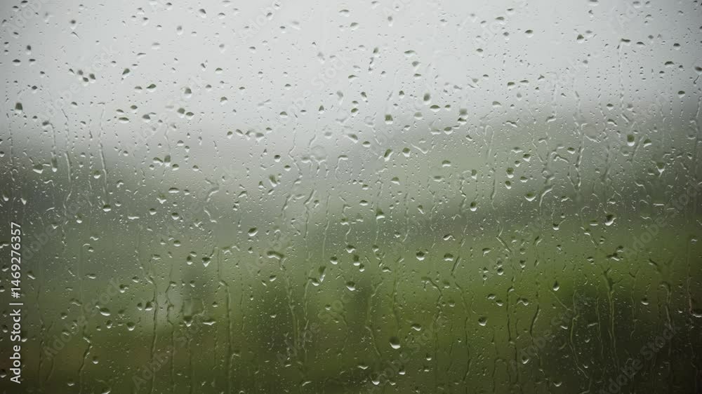 Close-up of raindrops streaming down a windowpane with a blurred natural background, capturing the calm and moody atmosphere of a rainy day.