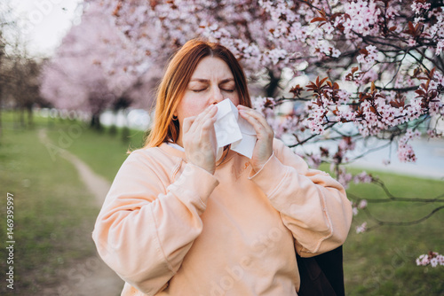 Sneezing young redhead woman with nose wiper among blooming trees in park. Portrait of sick women sneezes in white tissue, suffers from rhinitis and running nose. Symptoms of cold or allergy.