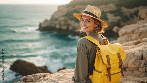 Fototapeta Naklejka Na Ścianę i Meble -  Smiling woman in a straw hat with a yellow backpack looks over her shoulder at the sea.