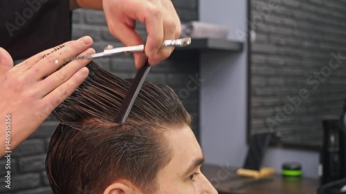 A professional hairdresser is cutting a young man's dark hair in a barber's shop using scissors and a comb.