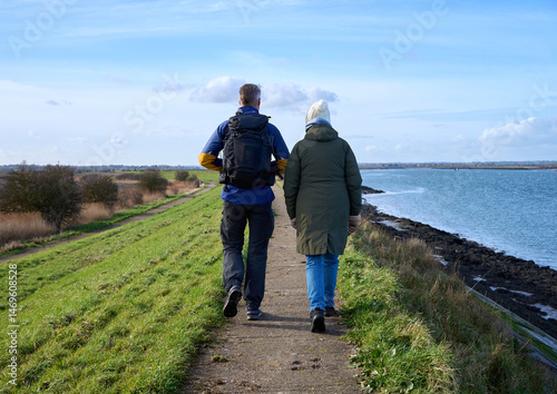 A senior mother and her adult son with a backpack walk along a scenic path by the water on a bright day.