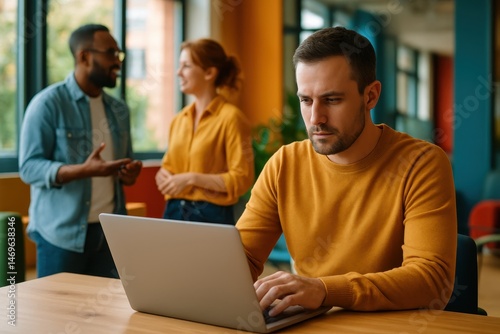 A man works on his laptop as a colleague engages with him in the background