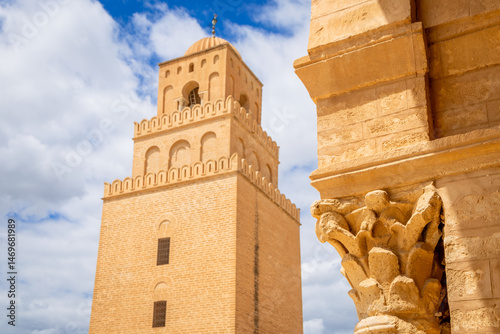 Old decorated column and view to minaret tower of the Great Mosque of Uqba with clouds in the blue sky, Kairouan, Tunis