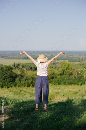 Senior woman enjoying freedom and nature, standing on a hill with arms raised. Back view of a carefree female in summer outfit and straw hat. Concept of relaxation, nature connection, and mindfulness.