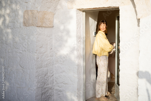 Canvastavla Smiling woman in yellow jacket opens a rustic door of a white trullo house