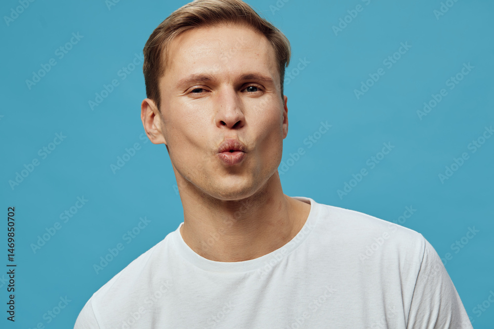 Fototapeta premium Confident young man with light brown hair making a playful kissy face, wearing a plain white t-shirt, shot against a bright blue background in a studio portrait, casual style, expression of fun and