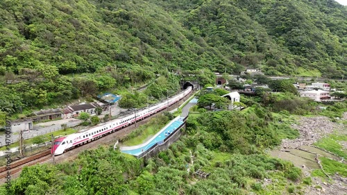 A sleek train glides along the coast of Shicheng, Yilan, between lush green hills and the rocky sea. The blue sky, ocean, and winding tracks create a peaceful, scenic blend of nature and travel.