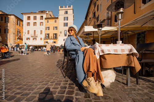 Canvas Print Smiling woman in a blue coat enjoys her coffee in a sunlit Roman square, surroun
