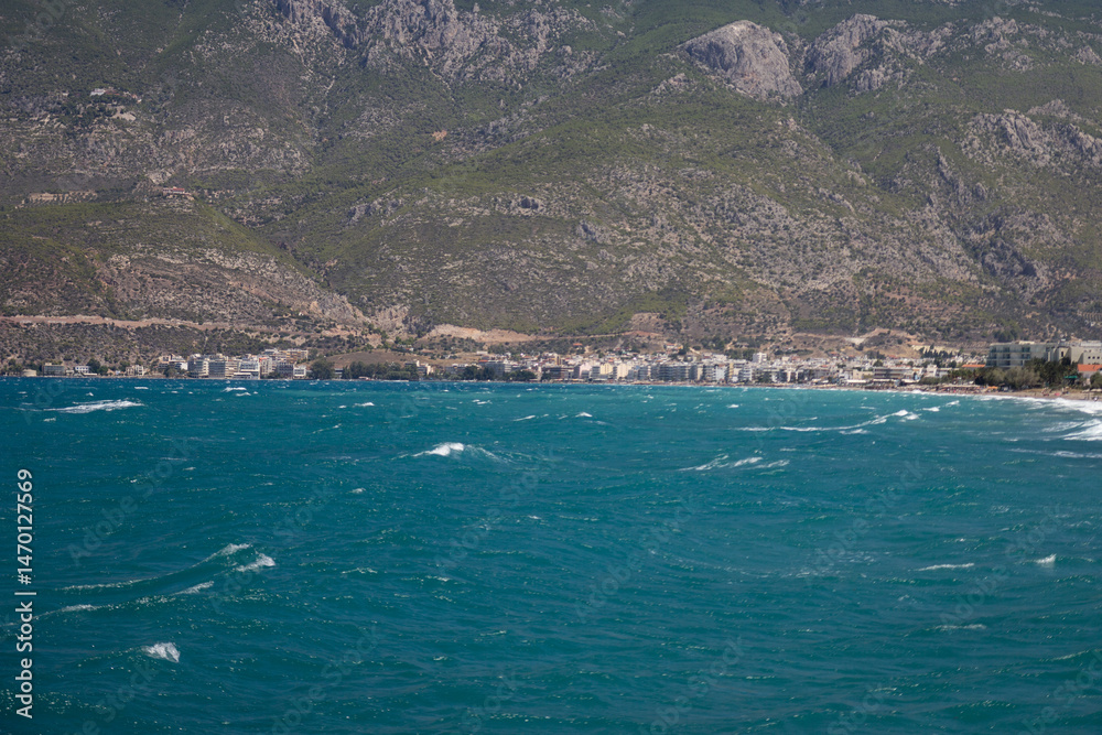 Obraz premium Crystal-clear blue waters near Loutraki sparkle under the sun, with a scenic mountain rising in the distance. A peaceful and inviting Mediterranean view ideal for travel and nature themes.
