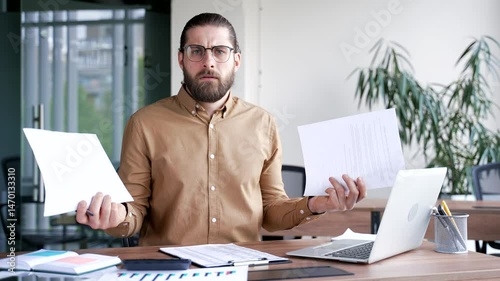 Confused businessman having difficulty with paperwork sitting at workplace in business office. Puzzled disappointed entrepreneur looks at documents, cannot understand the problem and looking at camera