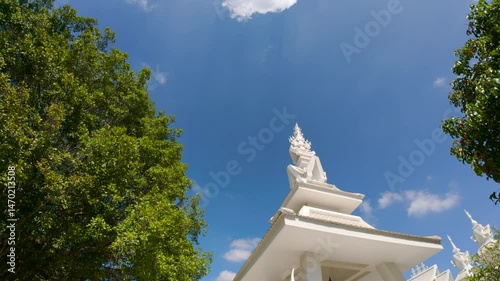 White temple spire beside green trees and blue sky at Wat Rong Khun in Chiang Rai, Thailand.
