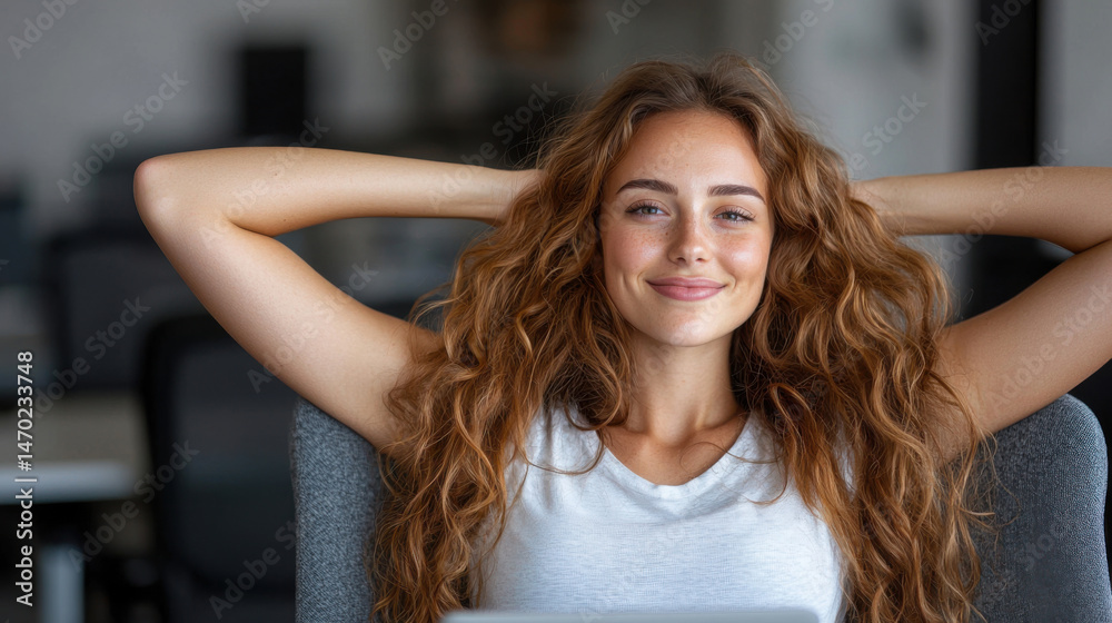 Fototapeta premium A woman with curly hair leans back in her chair, smiling confidently as she works on her laptop in a contemporary office setting filled with natural light