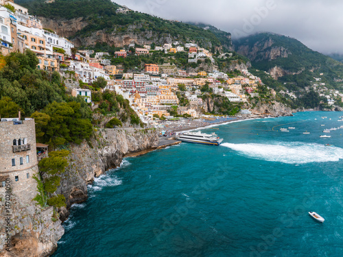 Fototapeta Naklejka Na Ścianę i Meble -  Coastal town of Positano, Italy, with stacked buildings on a steep hillside, a stone tower, turquoise waters, boats, and a cloudy sky.