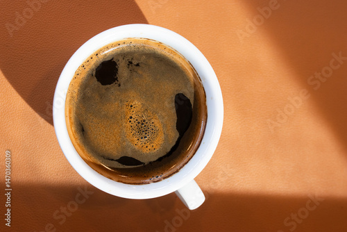A close-up of a freshly brewed black coffee in a white ceramic cup, sitting on a warm brown surface with natural light casting soft shadows.