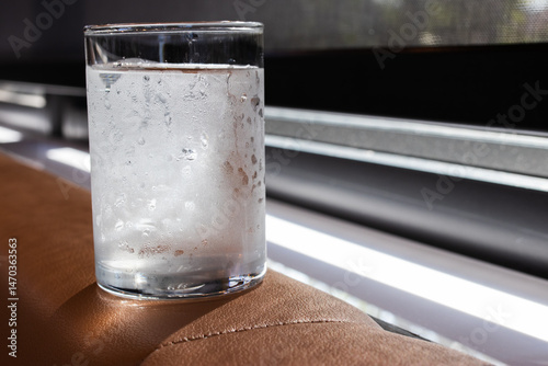 A cold glass of water with condensation sits on a brown surface by the window, catching natural light. The droplets suggest freshness and coolness, ideal for hydration or lifestyle themes.