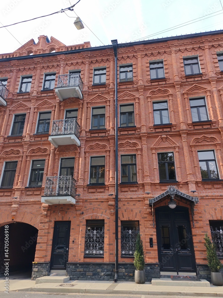 Fototapeta premium A tall red brick building featuring elaborate decorative elements, black iron balconies, and arched ground-floor entrances. The image reflects the grandeur and craftsmanship of vintage city