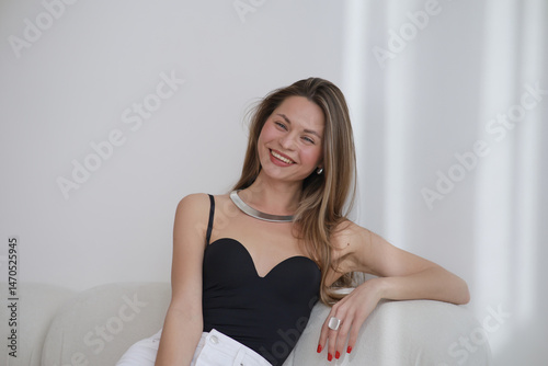 Portrait of a confident young woman with long straight hair and blue eyes, wearing a black strapless top and metallic necklace, posing indoors with soft natural lighting and a neutral background.