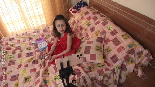 Little Girl in Red Dress Making a Video on Her Smartphone About Toy Nail Polish While Sitting on Her Bed
