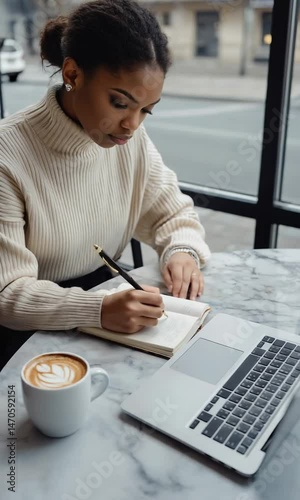 A student writing in a notebook on a caféStudent Writing in Notebook at Café Table with Coffee and Laptop Showing PDF E-Book, Natural Window Light, Blurred Street View, Overhead Composition with Righ