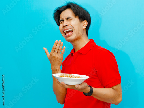 portrait of Asian man in a red polo shirt holding a plate of fried noodles showing a funny facial expression to spicy food on an isolated blue background.