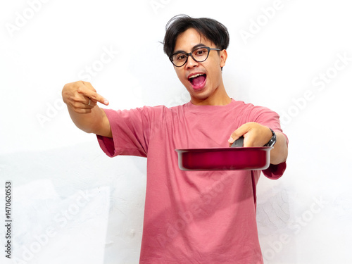 Young Asian man in a pink t-shirt excitedly points to a frypan while holding it. Isolated on a white background.