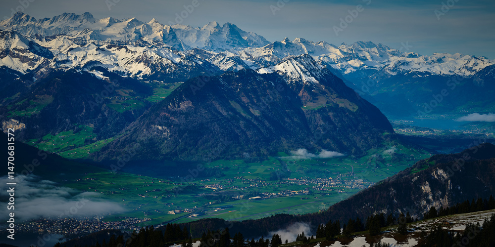 Naklejka premium Majestic snow-capped mountains tower above a serene green valley in Switzerland during a cloudy day