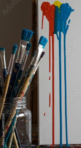 A vibrant still life featuring paintbrushes in a glass jar next to a canvas with streaks of red, yellow and blue paint dripping down. photo