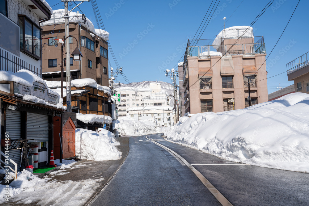 Fototapeta premium Winter street view in a snowy Japanese town with modern and traditional buildings. Tourists walk along cleared paths surrounded by high snowbanks, under a clear blue sky with mountain backdrop.