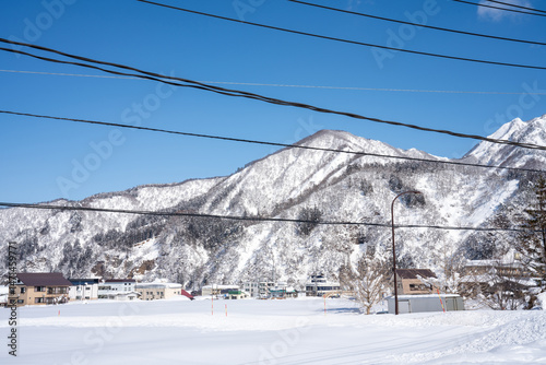 Snow-covered village nestled at the base of majestic winter mountains under a clear blue sky. Power lines cross the scene, adding contrast to the peaceful, frosty landscape.