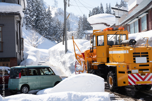 Bright orange snow removal vehicle parked on a snowy residential street in winter. The powerful machine stands out against the white snow and blue sky, highlighting essential infrastructure during