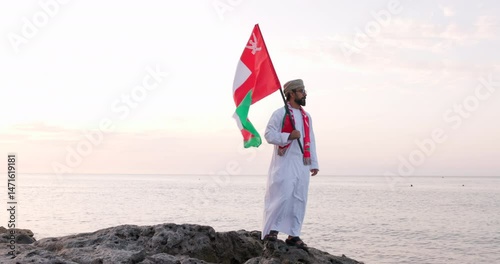 Omani man stands with Omani National Flag, wearing omani traditional dishdasha and Musaar