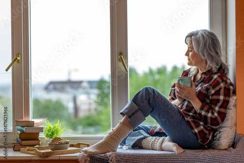 Fotomural An elderly woman sits on the windowsill with tea, books and thoughtfully looks o