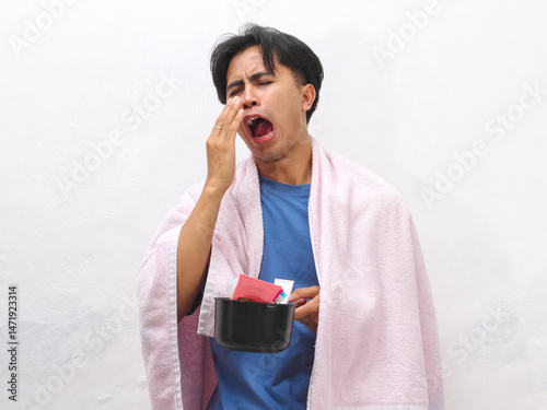 Portrait of a tired Asian man holding a towel and toiletries, appearing lazy and just awake, set against a white background. photo