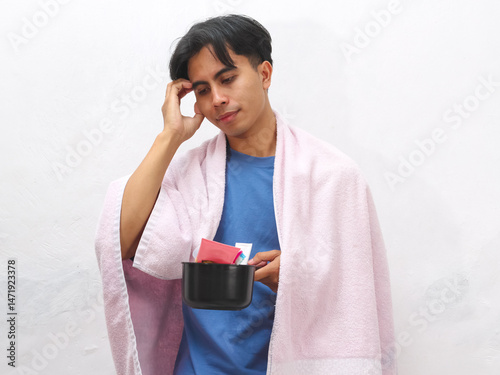 Portrait of a tired Asian man holding a towel and toiletries, appearing lazy and just awake, set against a white background. photo