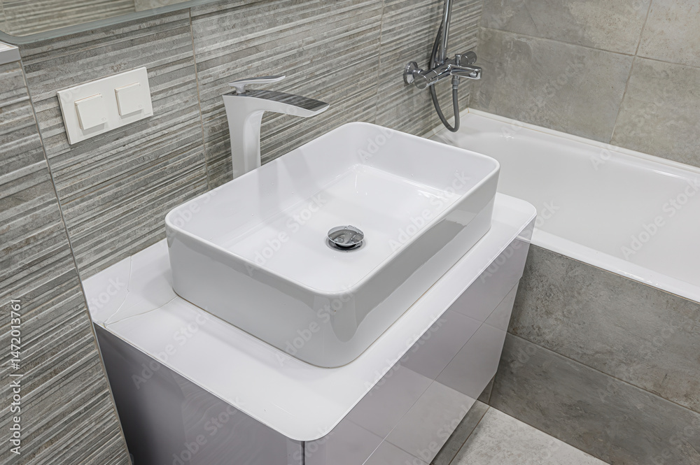 Fototapeta premium Modern bathroom scene featuring a rectangular white sink with a sleek faucet, set on a white cabinet against gray tiled walls. A bathtub is visible in the background