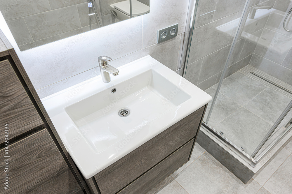 Fototapeta premium Modern bathroom featuring a white basin atop a wood-grain vanity, chrome faucet, and a tiled shower area.