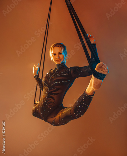 young woman in black costume is making aerial trick in longitudinal twine on the beige background in photo studio in warm and cold lights, sport concept, free space.