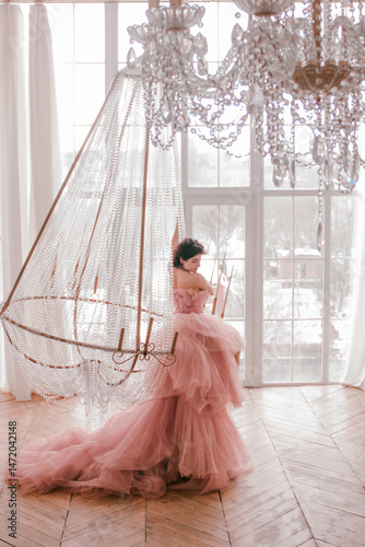 young woman with short black hair in pink princess dress with train is sitting on the crystal chandelier near huge panoramic window background luxury style. fashion concept, free space