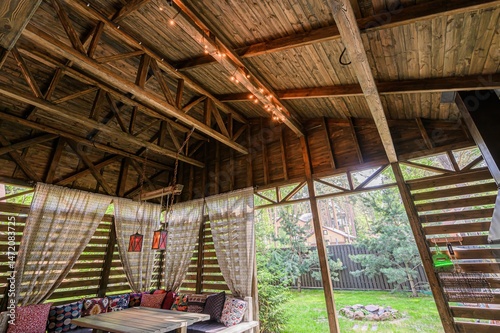 Rustic wooden gazebo with fabric curtains, colorful pillows, and string lights. View of trees outside