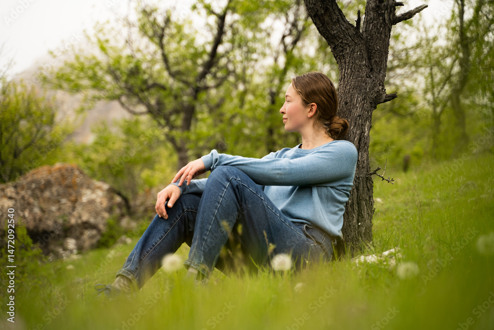 Young woman relaxing in spring forest, enjoying the silence, sitting on green grass under tree in daylight, wear blue sweater and jeans, peaceful nature moment, outdoor lifestyle. Side view full body Young woman relaxing in spring forest, enjoying the silence, sitting on green grass under tree in daylight, wear blue sweater and jeans, peaceful nature moment, outdoor lifestyle. Side view full body