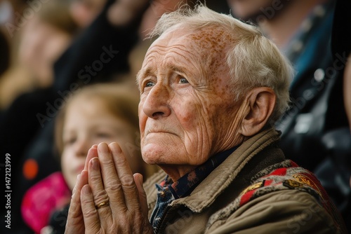 Elderly man with clasped hands looks upward in hope, faith, or concern in thoughtful reflection, surrounded by others at an event