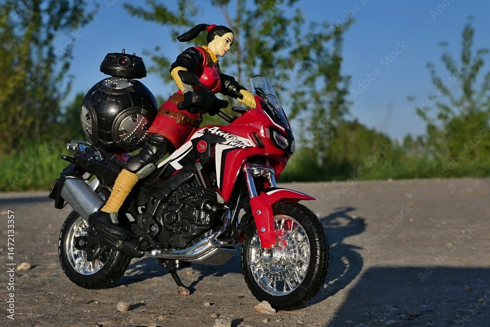 Naklejka premium GI JOE action figure of female intelligence agent Jinx riding on Honda Africa Twin motorbike, Star Wars imperial astromech BB-9e on rear seat. Spring afternoon sunshine.