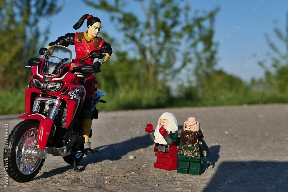 Obraz premium GI JOE action figure of female intelligentce agent Jinx, version 2004, standing with japanese dual sport motorbike Honda Africa Twin on asphalt road, greeting two LEGO dwarfes Balin and Dwalin on road