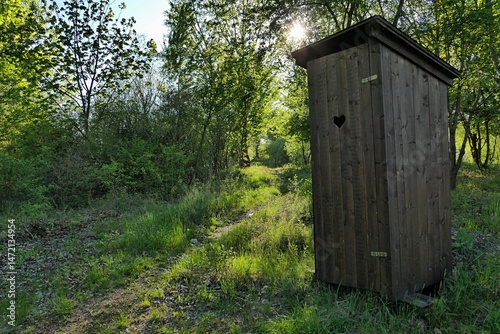 Fotografie Wooden latrine with typical hearth shaped small opening on doors, placed on pathway next to broadleaf trees surrounding fish pond