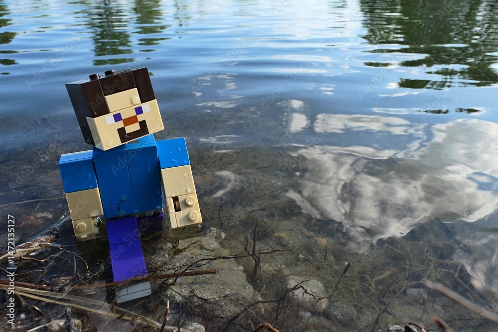 Naklejka premium LEGO Minecraft Steve walking out of shallow water of large spring fish pond, remains of aquatic plants under his feet and reflection of clouds visible.