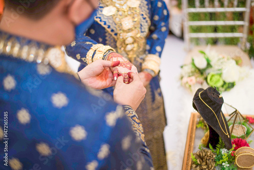 A happy bride and groom, wearing elegant Malay traditional attire during a ceremonial event, on a decorated stage during a wedding celebration. The groom is putting on the ring for the bride.