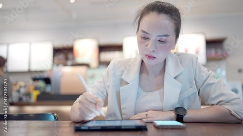 A woman is writing on a tablet with a pen. She is wearing a white jacket. A cell phone is on the table next to her