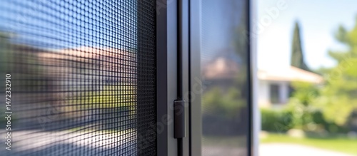 Close-up of window screen with blurred houses and greenery in sunlight.