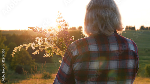 Lonely woman stands on hilly meadow in foothills on summer evening. She holds in hands bouquet of different blooming flowers collected at edge of forest in wild nature. Enjoying a warm sunset