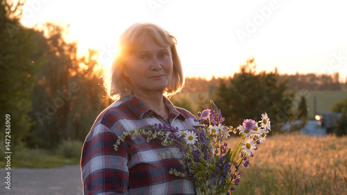 Portrait of elderly mature caucasian woman with bouquet of meadow flowers stands in countryside against sunset. Hobby is collecting flowers in wild. Tranquility and relaxation of quiet summer evening